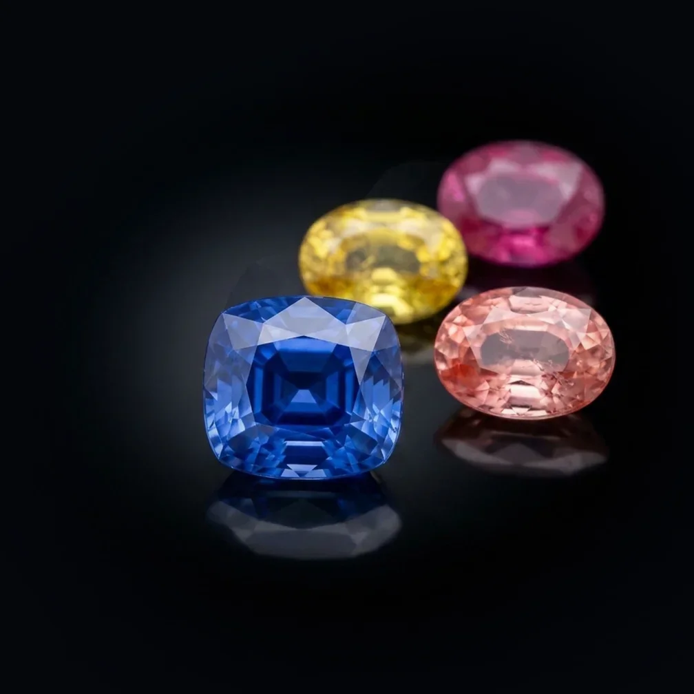 Four Ceylon stones on black: a blue cushion in front of a yellow oval, a pink oval and a padparadscha oval